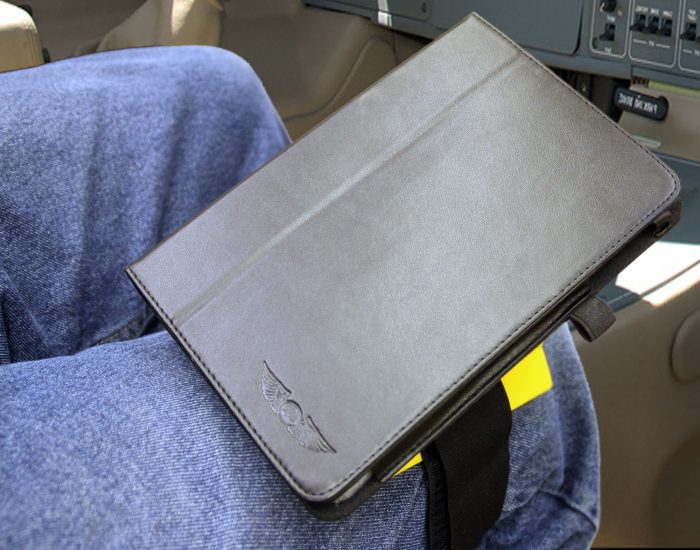 A closed black tablet case with a logo rests on a person's lap, who is wearing blue jeans. The background shows part of an airplane cockpit with visible controls.